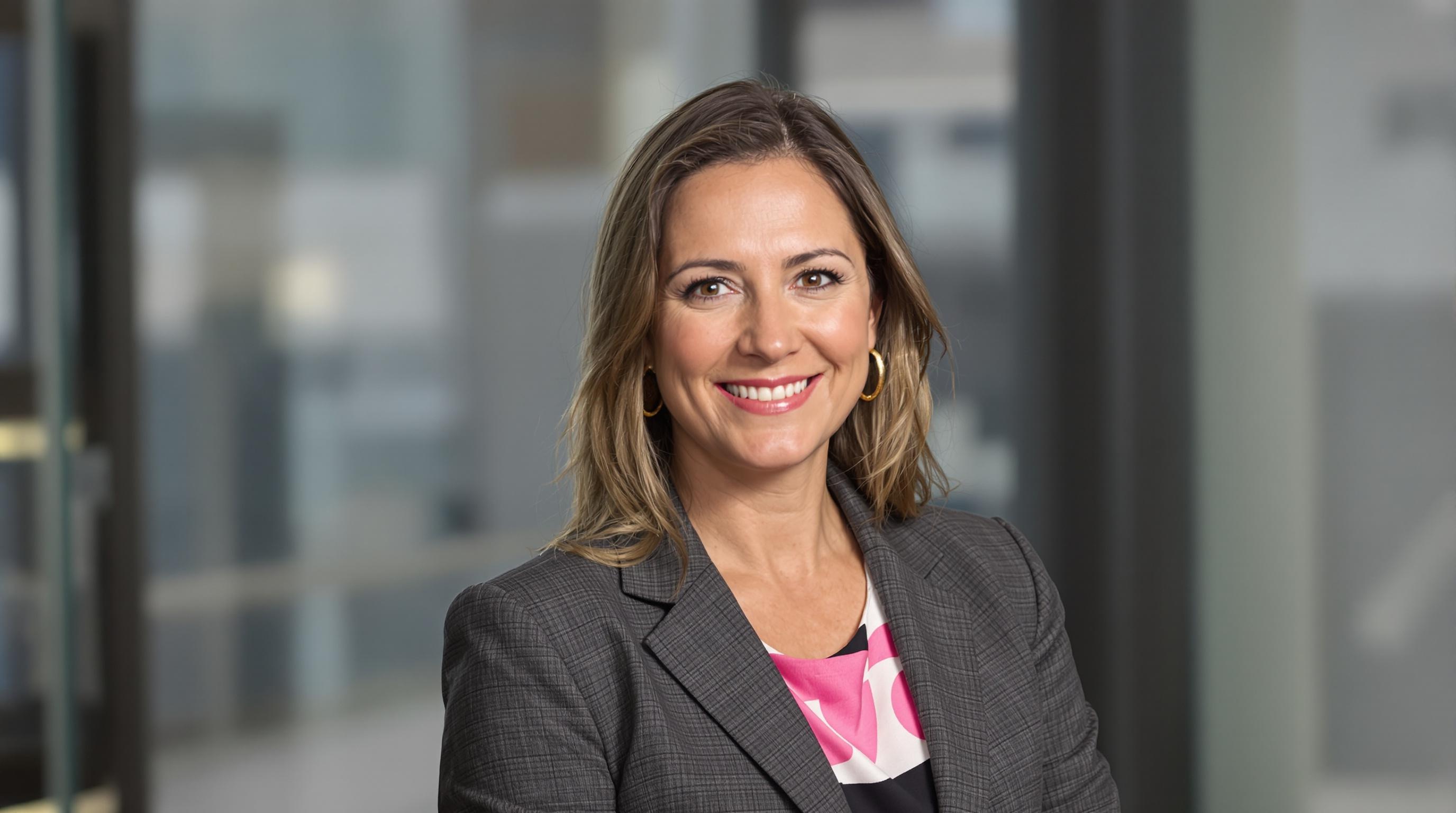 A middle-aged blond woman wearing a charcoal gray suit smiling and posing for a professional headshot in an office setting.